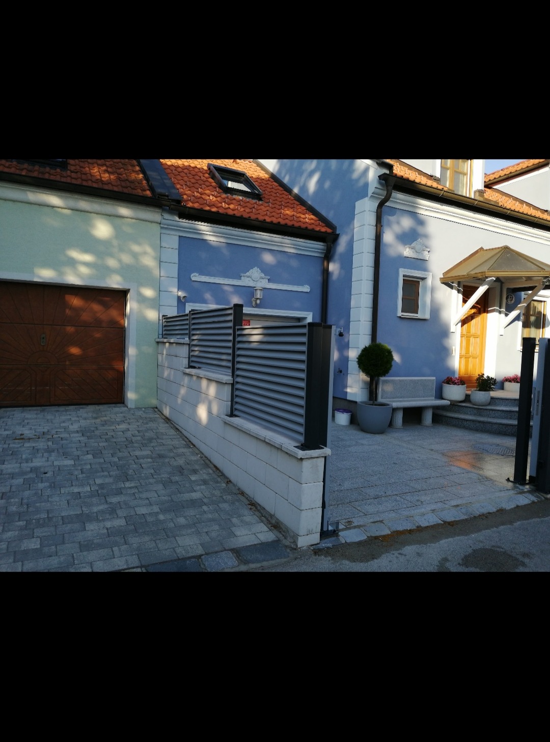 Nowoczesne, poziome ogrodzenie panelowe z szarych listew na niskim murku z betonowych bloczków, oddzielające podjazd z kostki brukowej od wejścia do domu z drewnianymi drzwiami i małą ławeczką.