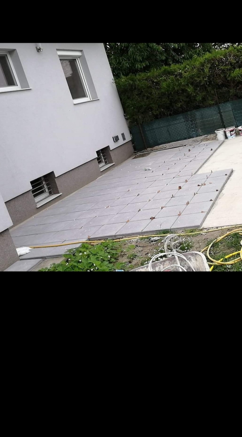 Układanie szarych płyt tarasowych na podłożu w Ludźmierzu, widoczne elementy poziomujące i fragment ogrodu zarośniętego roślinnością.