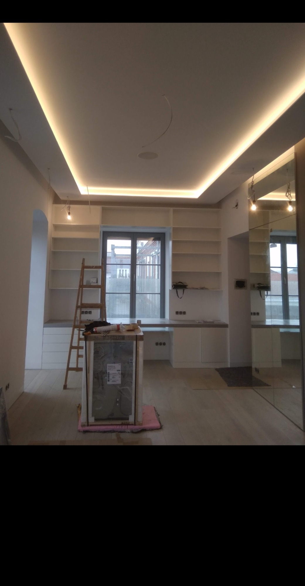 Jasne wnętrze w trakcie remontu z oknem, zabudowanymi półkami, drabina oparta o ścianę, podwieszany sufit z oświetleniem LED i zabezpieczony element na podłodze.