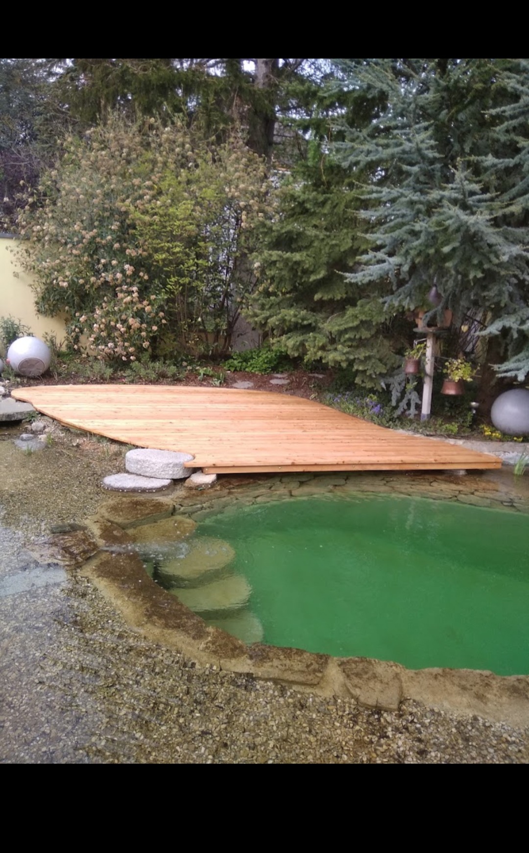 Drewniany taras nad stawem z zieloną wodą, otoczony kamieniami i roślinnością, w tle drzewa i krzewy.