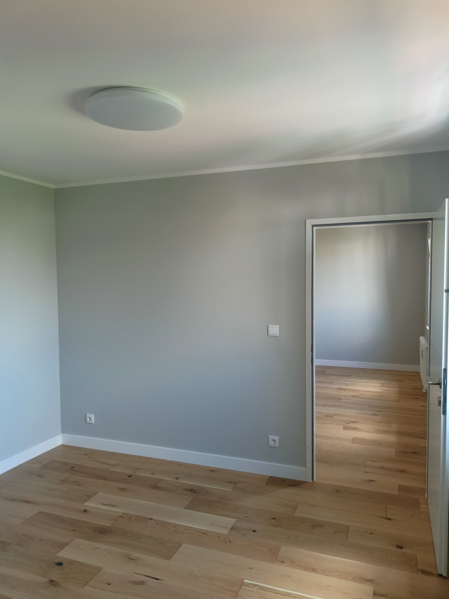 Jasne wnętrze po remoncie: świeżo pomalowane ściany w odcieniach szarości, drewniana podłoga i białe listwy przypodłogowe, widoczne gniazdka elektryczne i otwarte drzwi do kolejnego pomieszczenia.