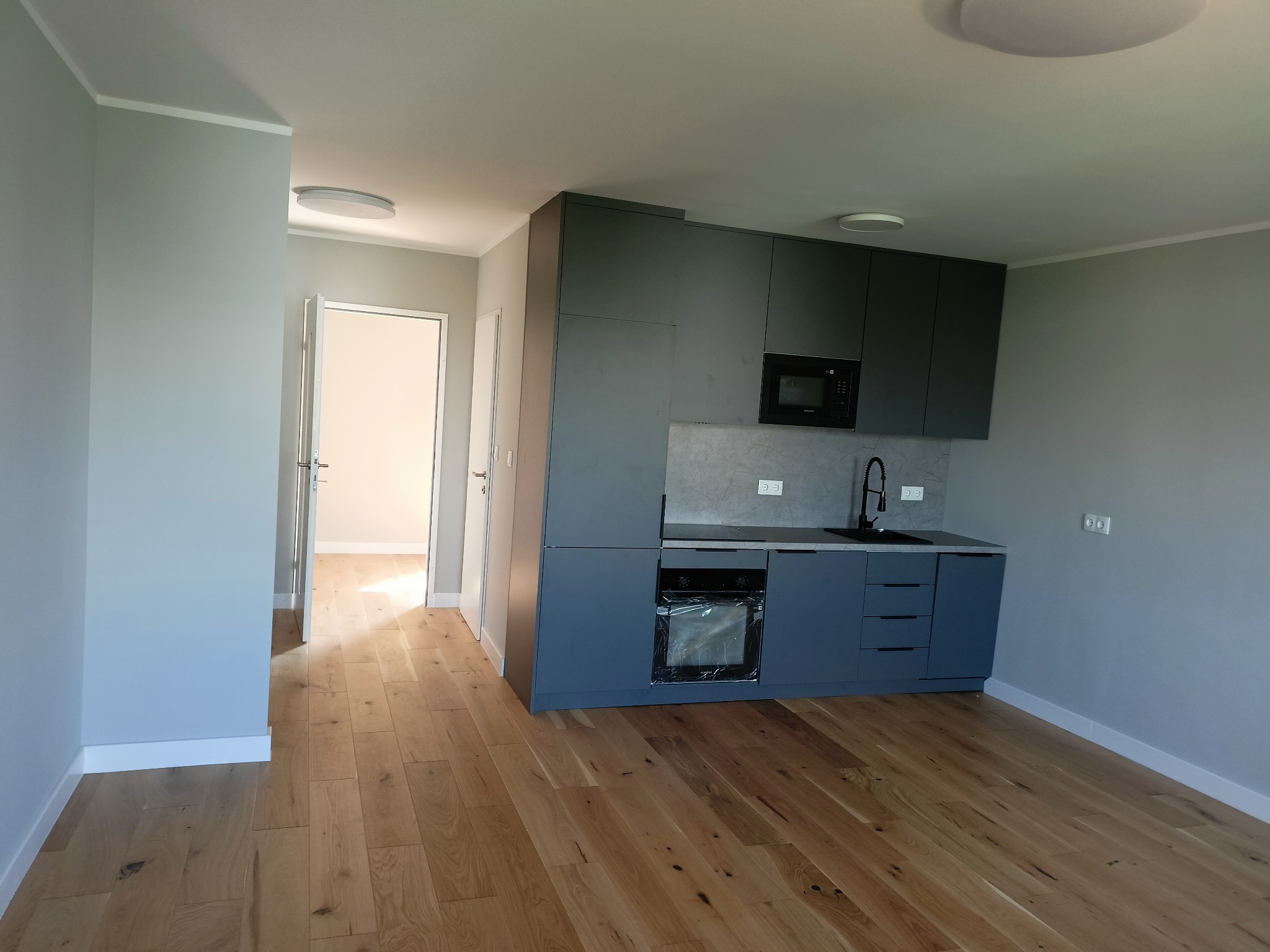 Wnętrze nowo wykończonego mieszkania z drewnianą podłogą i nowoczesną, szarą zabudową kuchenną z czarną armaturą i sprzętami AGD, widok na otwarte drzwi do kolejnego pomieszczenia.