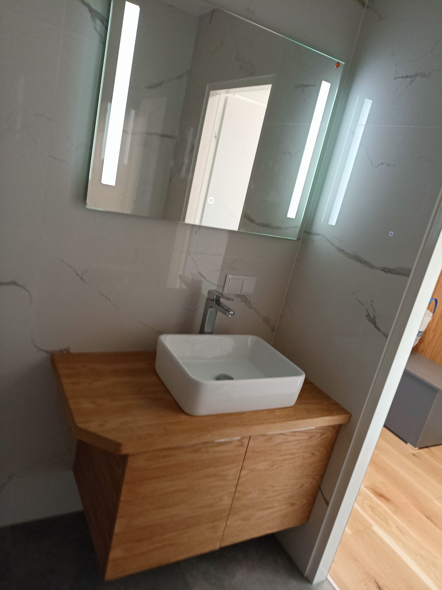 Nowoczesna łazienka z umywalką nablatową na drewnianej szafce, podświetlane lustro LED na ścianie z marmurowym wzorem.