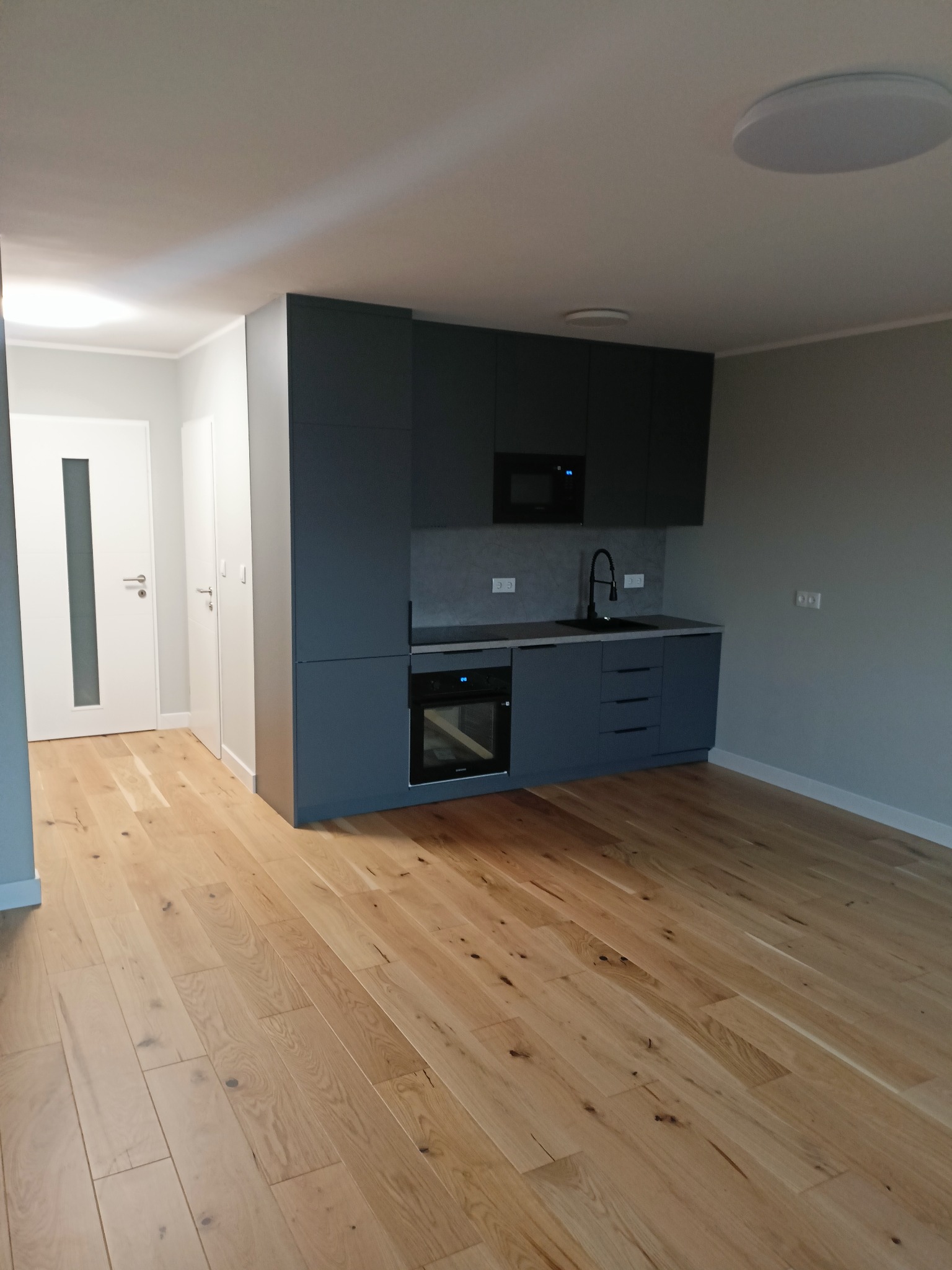 Nowoczesna, minimalistyczna kuchnia z szarymi szafkami, drewnianą podłogą i białymi drzwiami z przeszkleniem, widok z perspektywy wejścia.