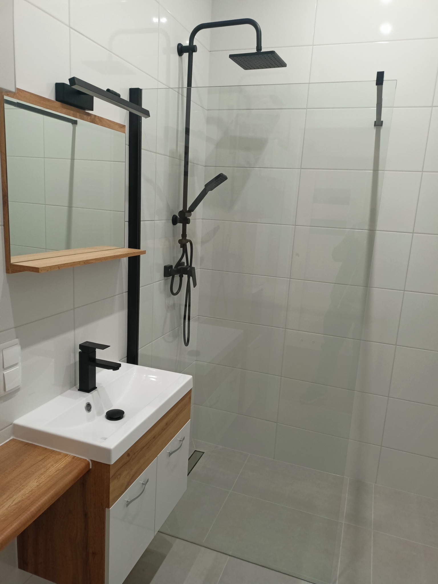 Nowoczesna łazienka z kabiną prysznicową oddzieloną szklaną ścianką, czarna armatura, biała umywalka z szafką z drewnianymi elementami i lustro z oświetleniem.