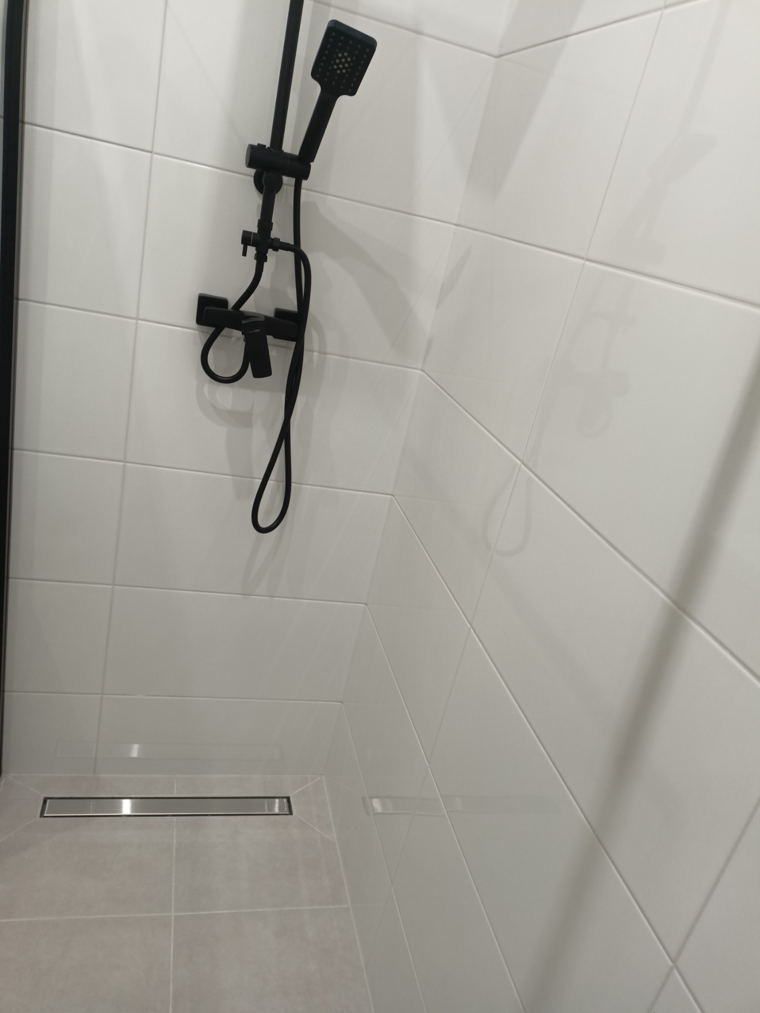 Nowoczesny prysznic z czarną armaturą i odpływem liniowym w jasnej łazience wyłożonej białymi i szarymi płytkami.