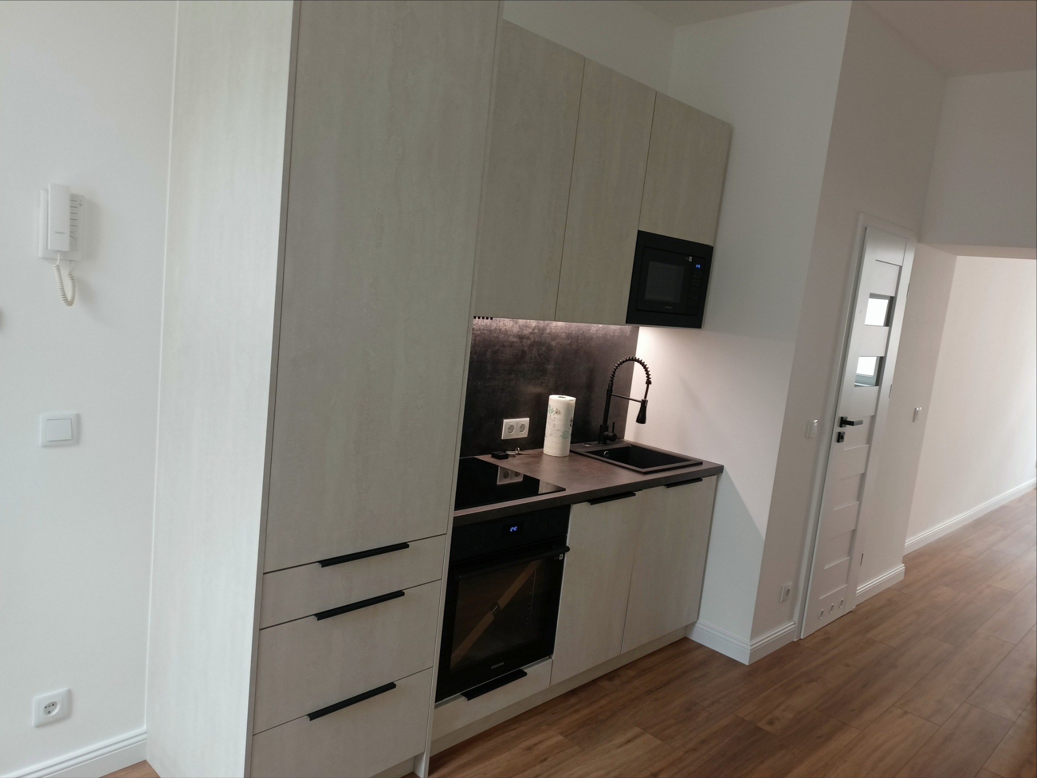 Nowoczesna, jasna kuchnia z beżowymi szafkami o fakturze kamienia, czarnymi uchwytami i sprzętem AGD, widoczna z perspektywy narożnej.