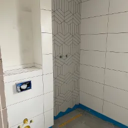 Remont łazienki w Pisz: Układanie płytek podłogowych i ściennych z geometrycznym wzorem, widoczne elementy instalacji sanitarnej oraz zabezpieczenia podłogi.
