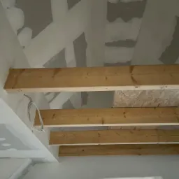 Wnętrze w trakcie budowy: sufit z drewnianymi belkami, częściowo pokryty płytami gipsowo-kartonowymi, widoczne przewody elektryczne i zabezpieczone okna folią.