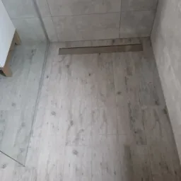 Ukończona łazienka z szarymi, drewnopodobnymi płytkami podłogowymi i odpływem liniowym, widok z góry, fragment kabiny prysznicowej po lewej.