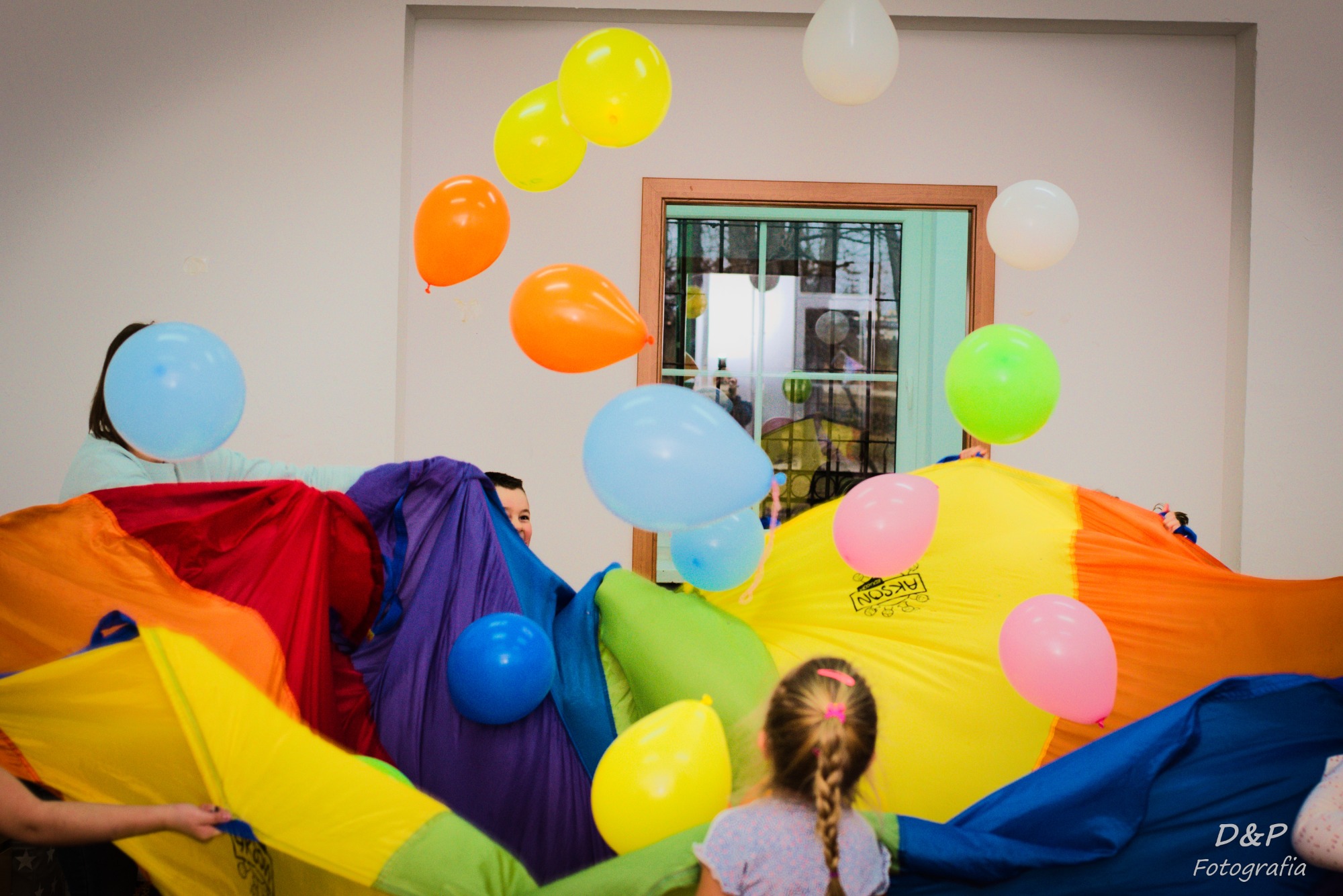 Dzieci bawiące się pod kolorową chustą animacyjną, unoszące balony w pomieszczeniu z oknem.
