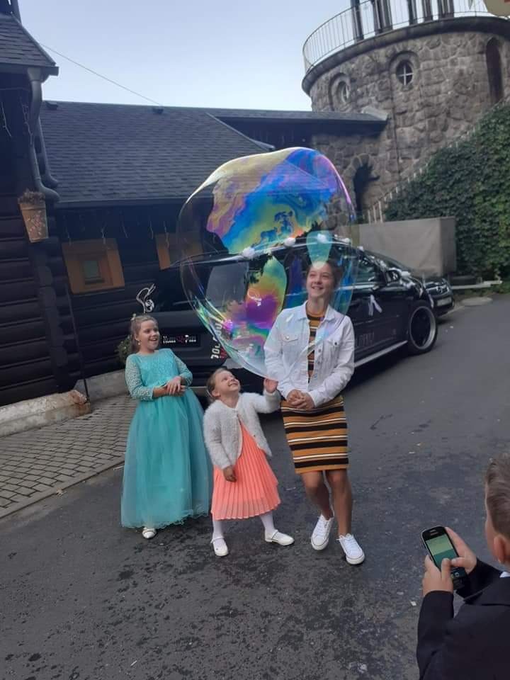 Trzy dziewczynki w sukienkach obserwują ogromną, tęczową bańkę mydlaną na tle czarnego samochodu i kamiennej wieży.