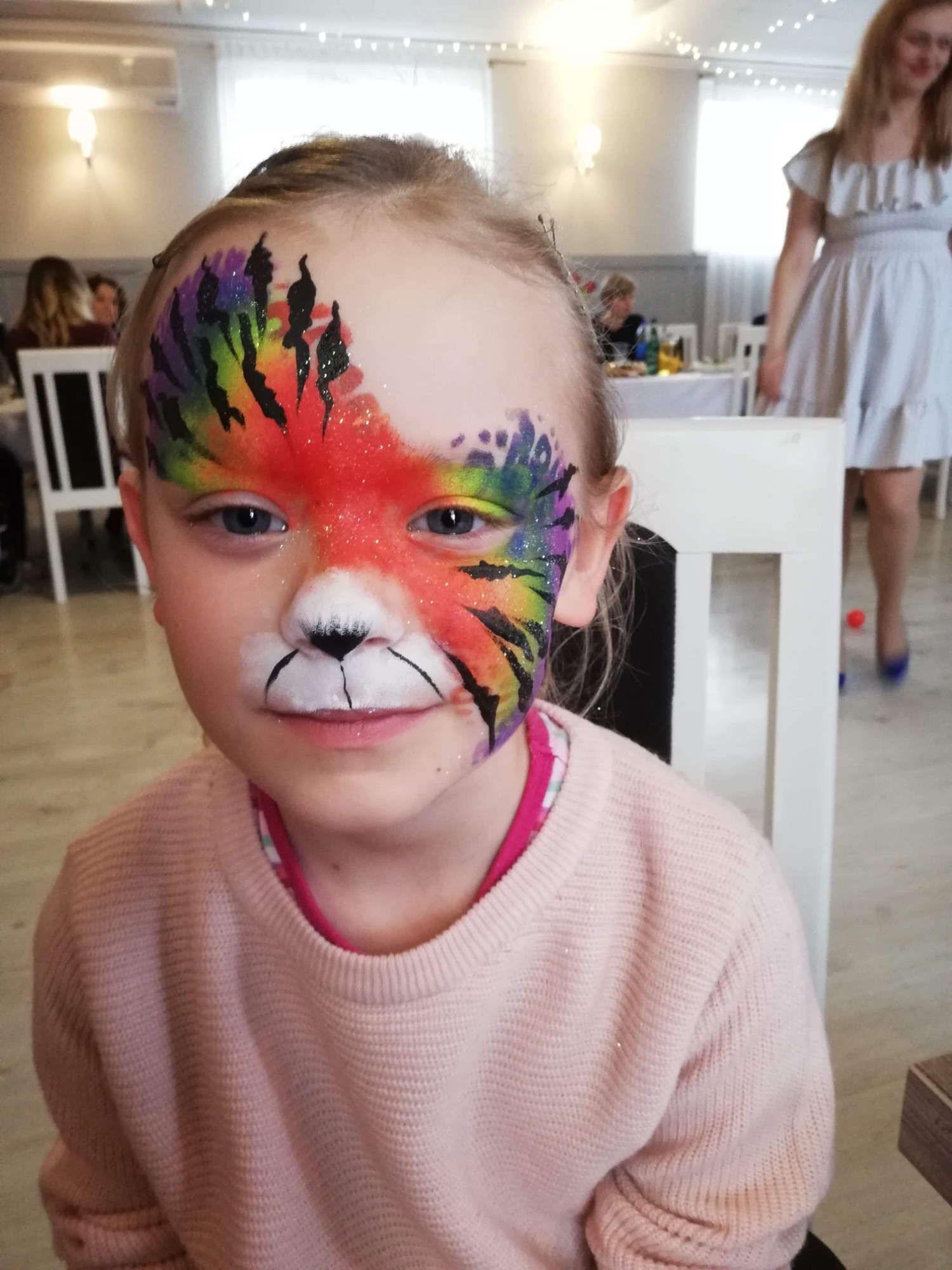 Dziewczynka z pomalowaną połową twarzy w motywy zwierzęce: czerwony lis z tęczowymi elementami i czarnymi paskami, podczas imprezy.