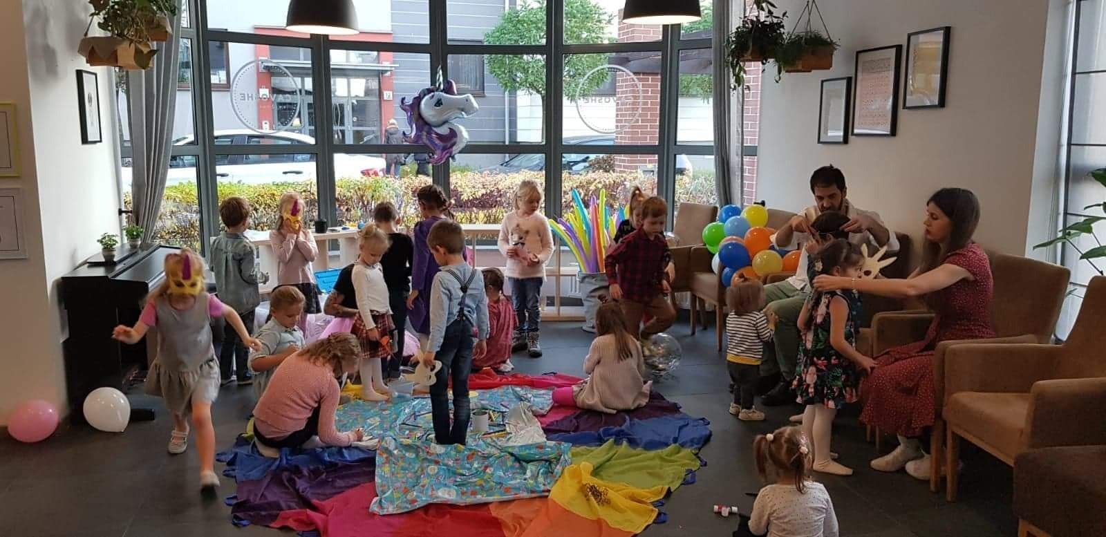 Grupa dzieci podczas zabawy z kolorowymi chustami i balonami w przestronnym pomieszczeniu z dużymi oknami; jedno dziecko biegnie w masce jednorożca, a dorosły zakłada maskę innemu dziecku.