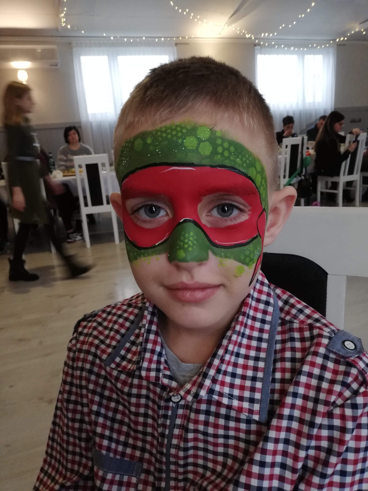 Portret uśmiechniętego chłopca z pomalowaną twarzą w stylu maski superbohatera (zielono-czerwona), w tle widoczni ludzie przy stolikach w sali.