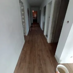 Korytarz w trakcie remontu z nową, drewnopodobną podłogą, surowe ościeżnice i wiadro z zaprawą w rogu.