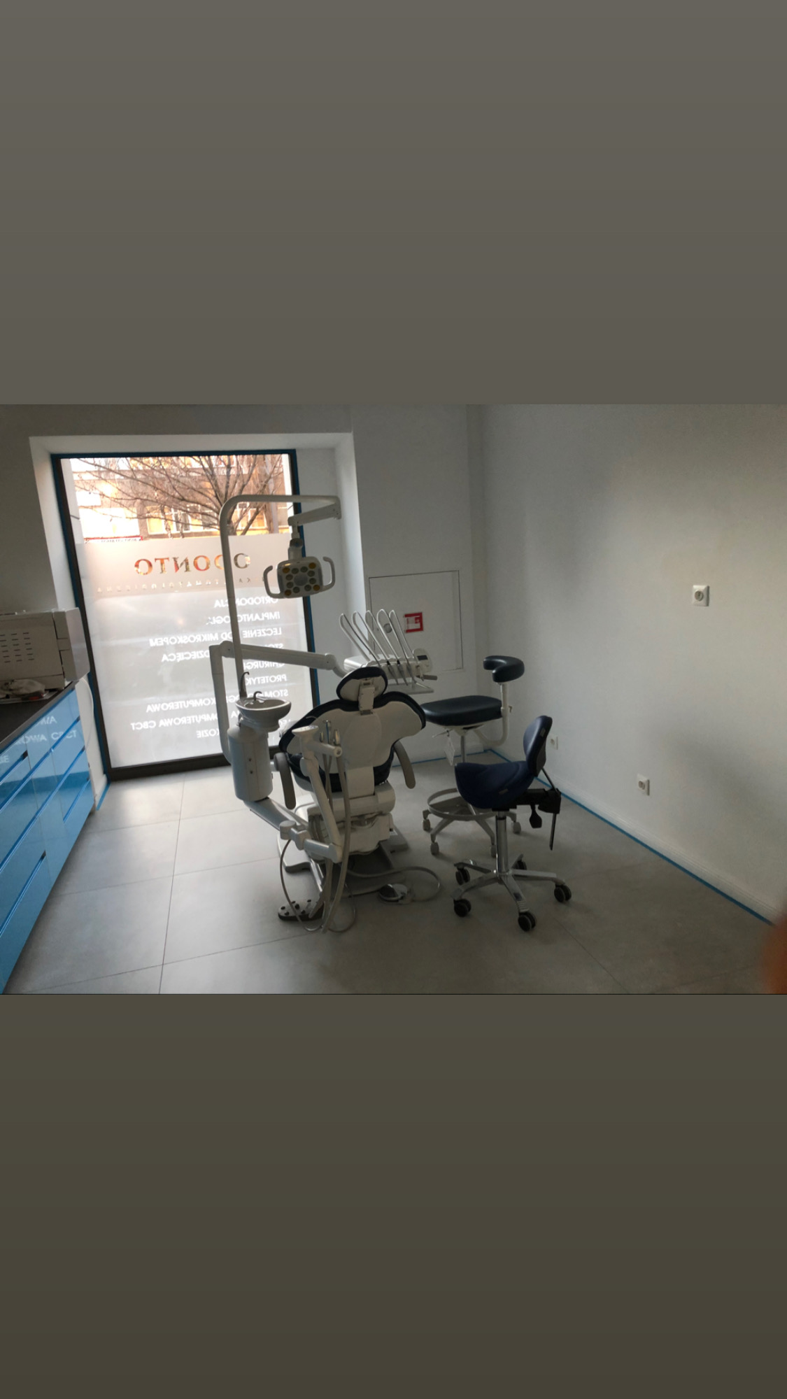 Wnętrze gabinetu stomatologicznego w Warszawie po remoncie, widoczne krzesło dentystyczne, lampa, szafki z niebieskimi frontami i duże okno z widokiem na drzewa.