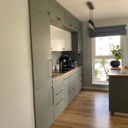 Nowoczesna kuchnia w odcieniach zieleni i bieli z drewnianym blatem, oświetlona lampą wiszącą z czarnym abażurem i naturalnym światłem z okna z szarą roletą rzymską, uzupełniona rośliną doniczkową...