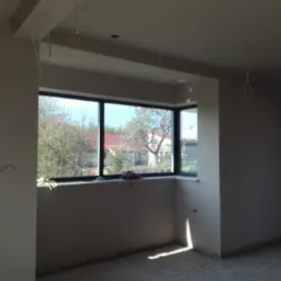 Wnętrze w stanie surowym po nałożeniu tynk&oacute;w, widoczne okno z widokiem na drzewa i dom z czerwoną dach&oacute;wką, na parapecie leżą drobne przedmioty.