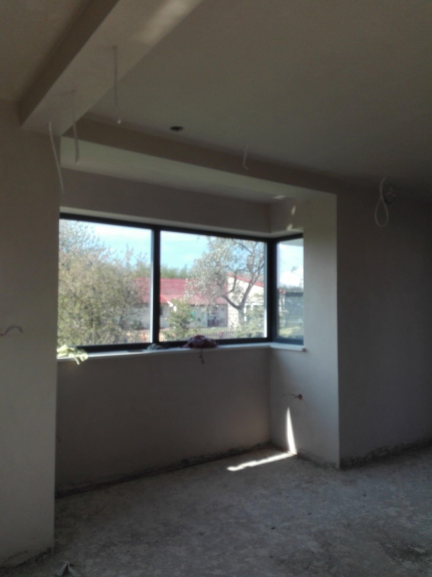 Wnętrze w stanie surowym po nałożeniu tynków, widoczne okno z widokiem na drzewa i dom z czerwoną dachówką, na parapecie leżą drobne przedmioty.