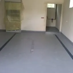 Szara, gładka podłoga z ciemnymi liniami wzdłuż ścian w pustym pomieszczeniu z białymi ścianami. Widoczne instalacje hydrauliczne i drzwi do innego pomieszczenia.