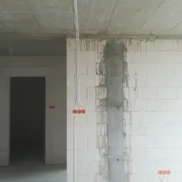 Surowy stan wnętrza z widoczną instalacją elektryczną: białe kable prowadzone po ścianach z betonu komórkowego i suficie, puszki elektryczne w kolorze czerwonym, betonowy filar z zaciekami cementu.