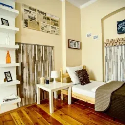 Pokój hostelowy z łóżkiem pojedynczym, drewnianym zagłówkiem ozdobionym plasterkami drewna, białą pościelą i brązowym pledem, obok biały stolik nocny z lampką, na ścianie tablica z napisem...