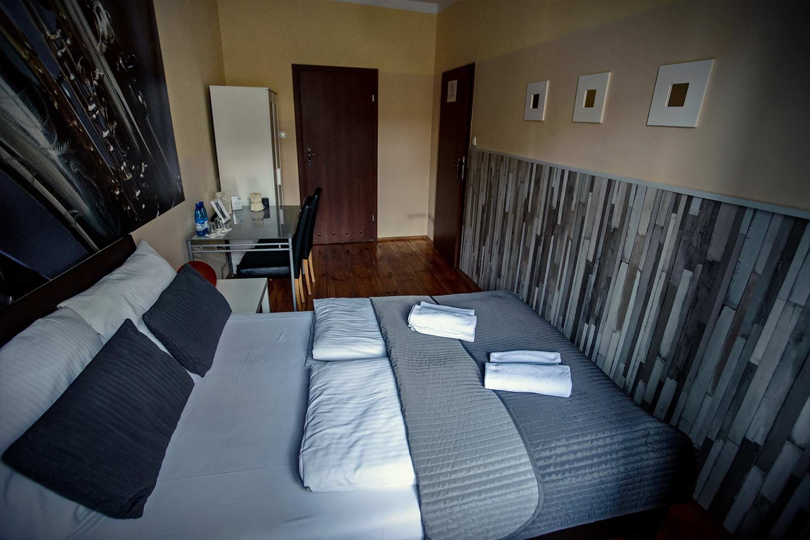 Pokój hotelowy z łóżkiem, biurkiem i dekoracyjną ścianą z drewnianych paneli w odcieniach szarości i brązu, nad łóżkiem wiszą trzy małe obrazy w białych ramkach.