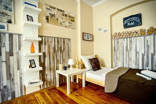 Pokój hostelowy z łóżkiem pojedynczym, drewnianym zagłówkiem ozdobionym plasterkami drewna, białą pościelą i brązowym pledem, obok biały stolik nocny z lampką, na ścianie tablica z napisem...