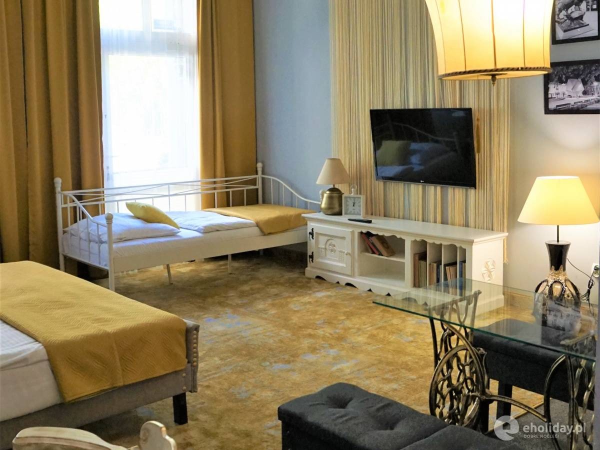 Pokój hotelowy z dwoma łóżkami pojedynczymi, telewizorem na ścianie i stolikiem z blatem szklanym na podstawie maszyny do szycia Singer.