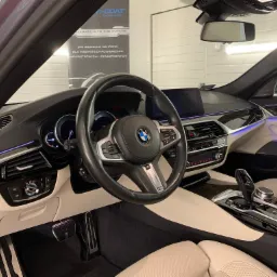 Wnętrze luksusowego samochodu BMW serii 5 po detailingu, z kremową sk&oacute;rzaną tapicerką, czarną deską rozdzielczą i kierownicą, oraz podświetleniem LED. Widoczny plakat reklamowy firmy detailingowej...