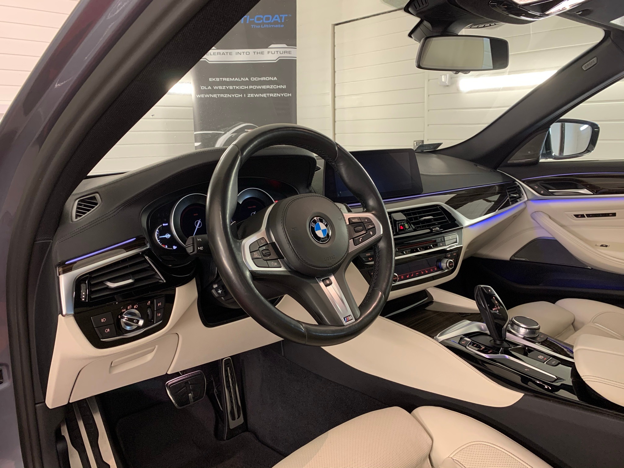 Wnętrze luksusowego samochodu BMW serii 5 po detailingu, z kremową skórzaną tapicerką, czarną deską rozdzielczą i kierownicą, oraz podświetleniem LED. Widoczny plakat reklamowy firmy detailingowej...
