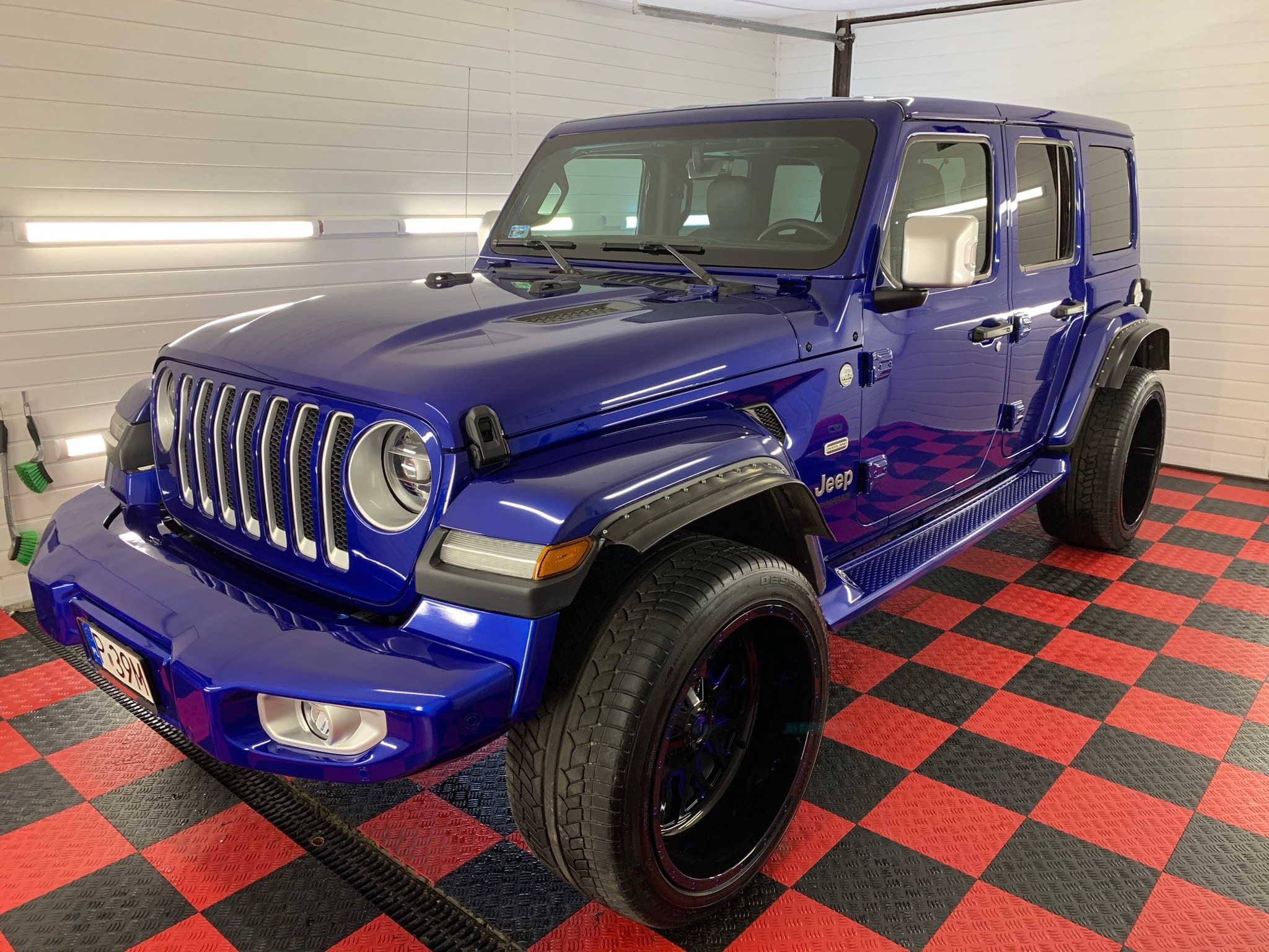 Błyszczący, granatowy Jeep Wrangler po umyciu, z widoczną strukturą opon i odbiciem światła w karoserii, na podłodze w czarno-czerwoną szachownicę.
