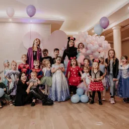 Grupa dzieci w kostiumach, pozujących z animatorkami przebranymi za postacie z bajek, na tle dekoracji z balonów.