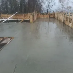 Świeżo wylany fundament z drewnianym szalunkiem w pochmurny dzień. Widoczne drzewa w tle.