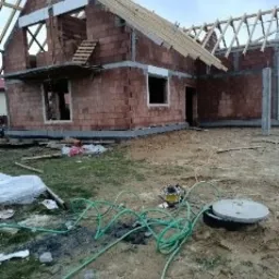 Budowa domu jednorodzinnego z czerwonej cegły w Niedźwiadzie, widoczne ściany w trakcie murowania, więźba dachowa w budowie, niezagospodarowany teren wokół.