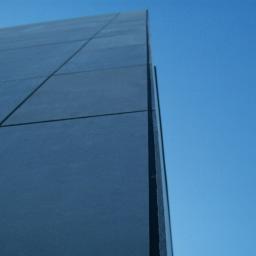 Lewandowski - Fragment elewacji budynku obłożonej ciemnymi płytami, widoczne regularne linie podziału między płytami na tle błękitnego nieba.