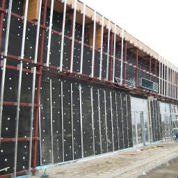 Lewandowski - Montaż elewacji budynku komercyjnego: widoczna warstwa izolacji z czarnej wełny mineralnej z białymi kołkami, metalowa konstrukcja nośna, pracownicy na drabinie i na ziemi, przygotowujący się...