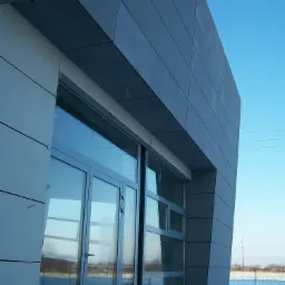 Nowoczesna elewacja budynku z szarych paneli kompozytowych i aluminiową stolarką okienną, widoczna z dołu pod kątem, z elementami rusztowania u góry.