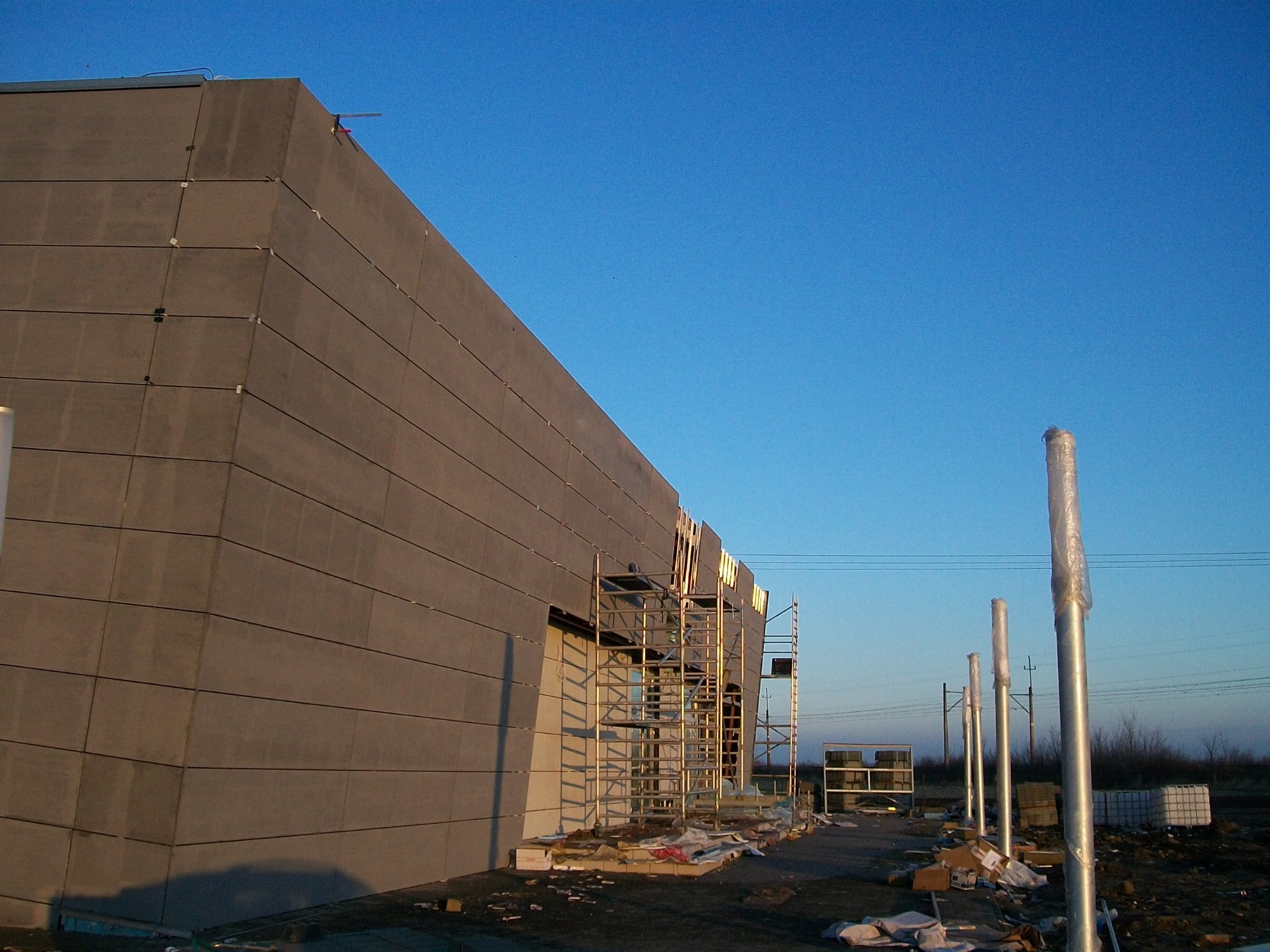 Elewacja budynku przemysłowego z szarego betonu w trakcie montażu, widoczne rusztowania i przygotowane słupy oświetleniowe z folią ochronną, błękitne niebo.