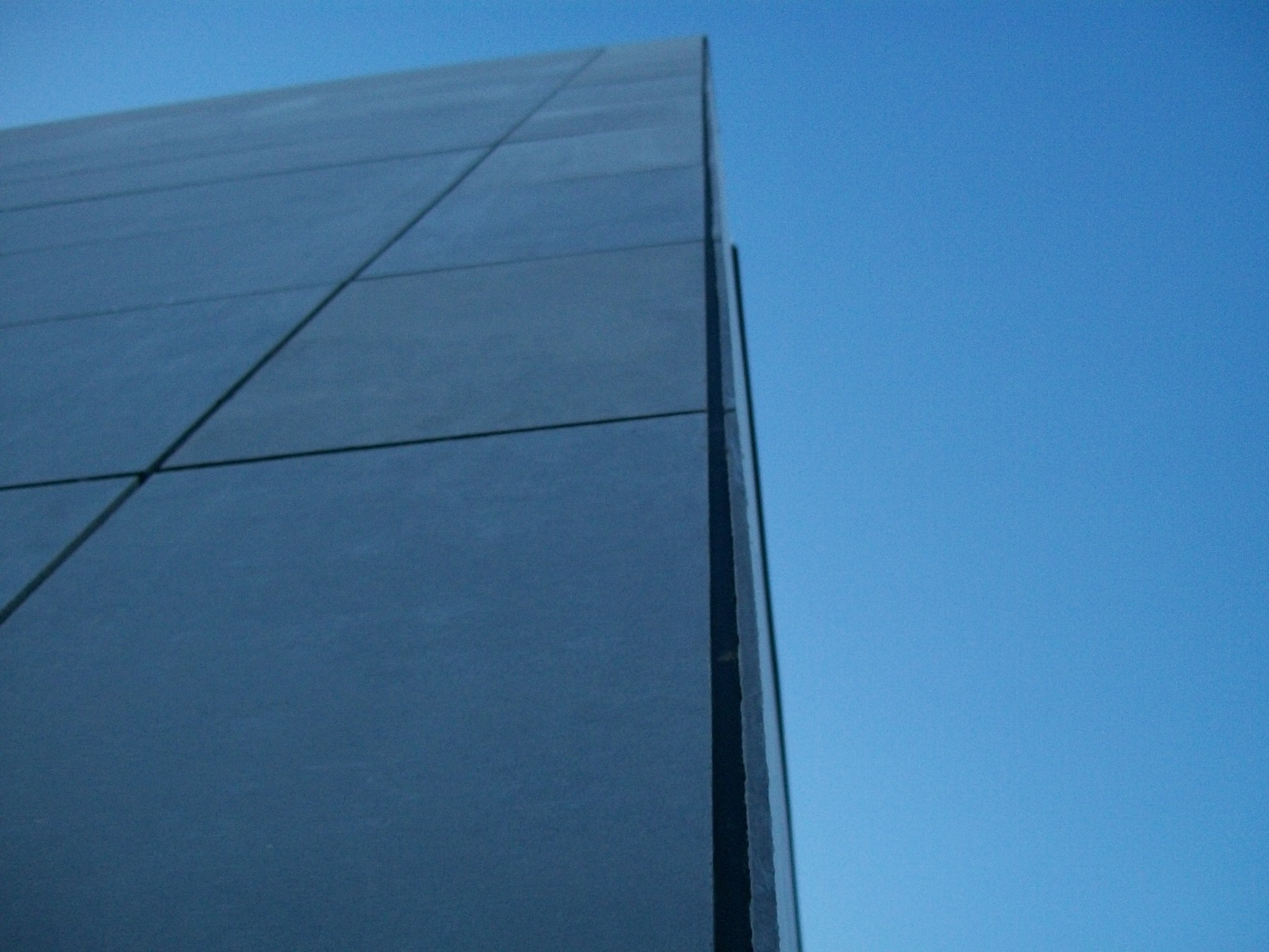 Fragment elewacji budynku obłożonej ciemnymi płytami, widoczne regularne linie podziału między płytami na tle błękitnego nieba.