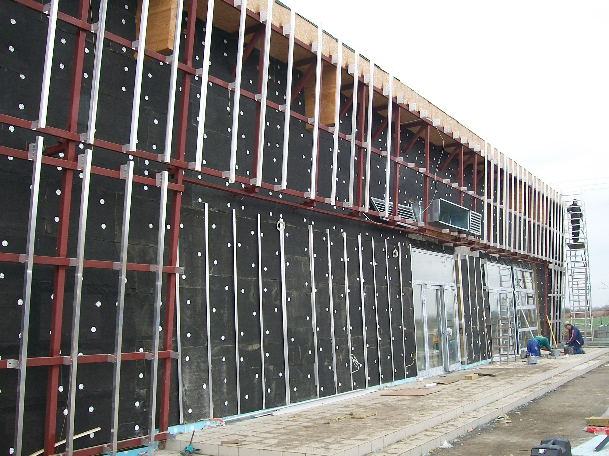 Montaż elewacji budynku komercyjnego: widoczna warstwa izolacji z czarnej wełny mineralnej z białymi kołkami, metalowa konstrukcja nośna, pracownicy na drabinie i na ziemi, przygotowujący się...