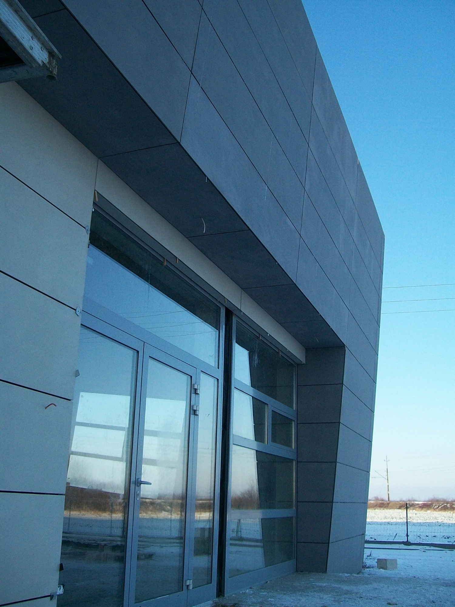 Nowoczesna elewacja budynku z szarych paneli kompozytowych i aluminiową stolarką okienną, widoczna z dołu pod kątem, z elementami rusztowania u góry.