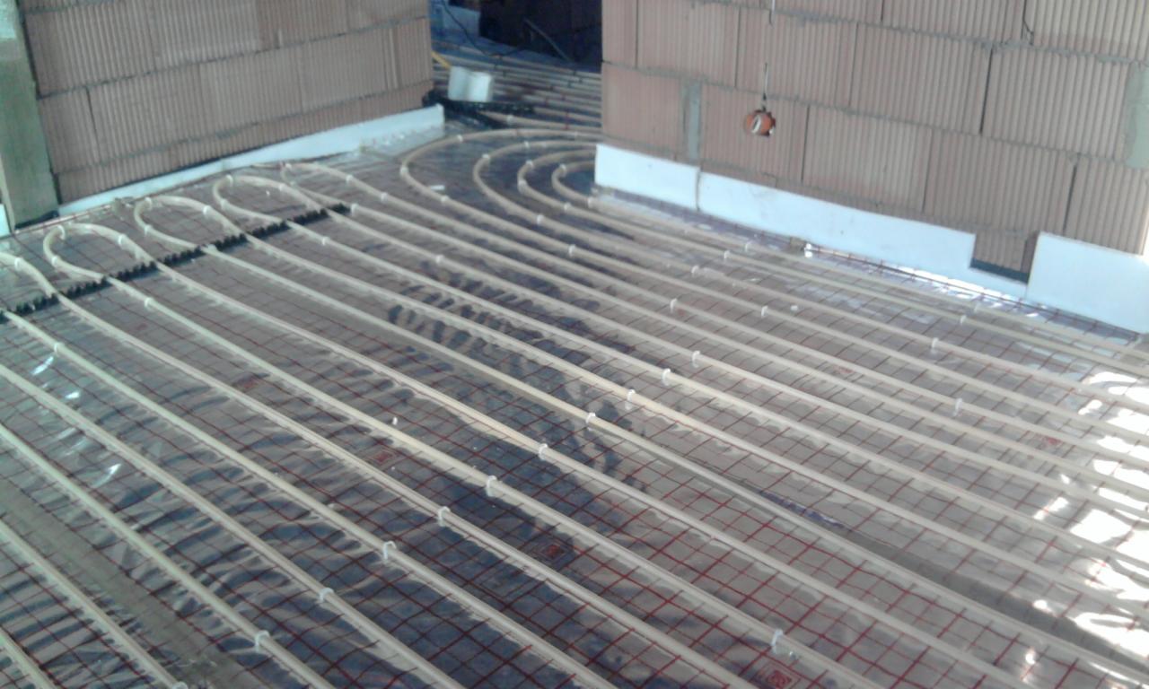 Instalacja ogrzewania podłogowego w trakcie montażu, widoczne rury grzewcze ułożone na folii z siatką, ściany z pustaków ceramicznych w tle.