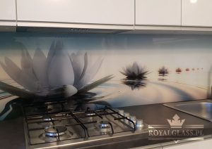 Szklana ściana nad blatem kuchennym z motywem kwiatów lotosu odbijających się w wodzie, zamontowana między białymi szafkami, widoczna płyta gazowa i zlewozmywak.