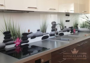 Szklany panel kuchenny z motywem zen: ułożone kamienie, trawy i kwiat lotosu, zamontowany nad blatem kuchennym ze zlewozmywakiem i płytą indukcyjną. Widoczne białe szafki wiszące.