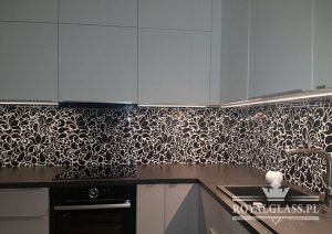 Szklany panel kuchenny z czarno-białym, kwiatowym wzorem, zamontowany między blatem a szafkami, oświetlony listwą LED. Widoczny fragment zlewu i płyty indukcyjnej.