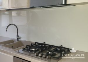 Beżowy panel szklany nad kuchenką gazową w nowoczesnej kuchni, widoczny kran i zlewozmywak, logo firmy w prawym dolnym rogu.