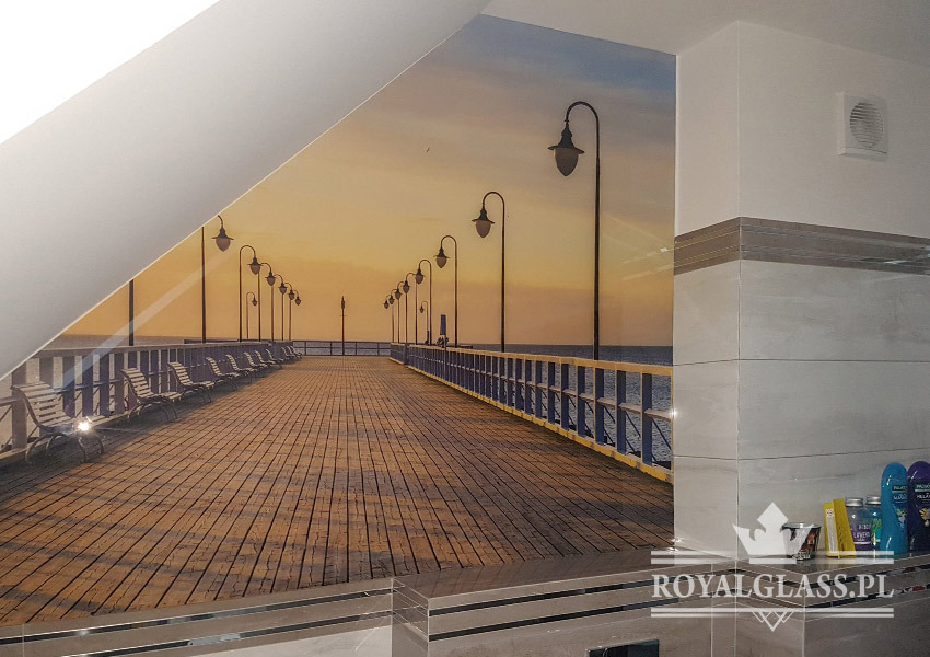 Szklana ściana z nadrukowanym motywem molo o zachodzie słońca, zamontowana w łazience, widoczny fragment umywalki i logo firmy RoyalGlass.pl w prawym dolnym rogu.