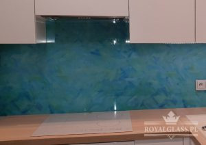 Szklany panel ścienny w kuchni, utrzymany w odcieniach błękitu i zieleni, imitujący abstrakcyjny wzór akwareli, zamontowany między blatem a szafkami, z widocznym gniazdkiem elektrycznym po prawej...